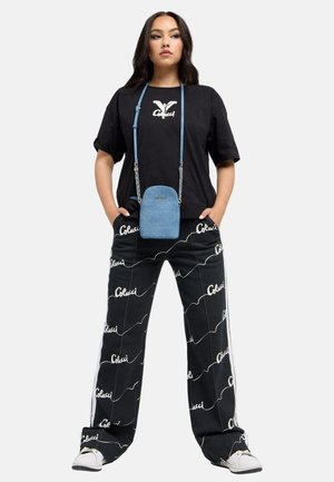 Vrouw staat met handen in de zakken, draagt een zwart T-shirt met het Colucci-logo, zwarte broek met witte Colucci-tekst, witte sneakers en een kleine blauwe crossbodytas.