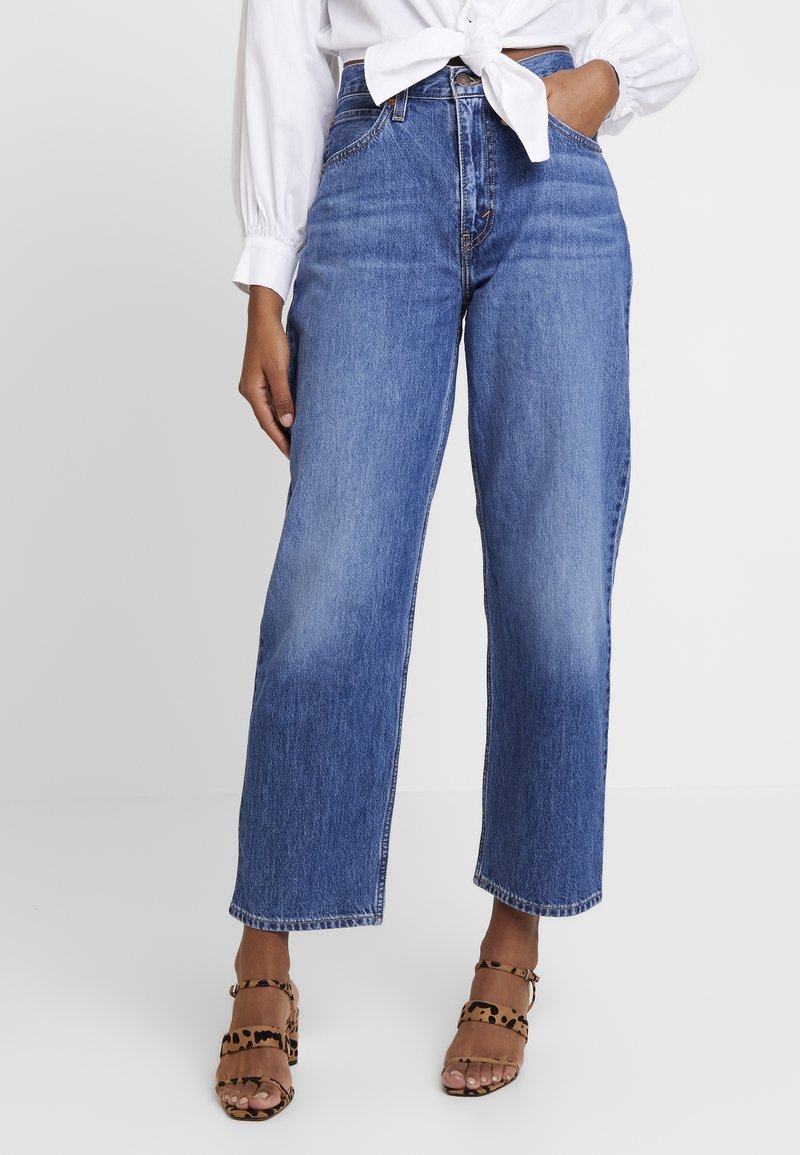 Femme portant un jean bleu taille haute à jambes droites, un chemisier blanc à manches longues noué, et des talons ouverts à imprimé léopard.