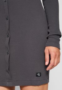 Grå ribbad klänning med långa ärmar, knapplångd och en CK-logotyp patch nära fållen. Tyget verkar mjukt och åtsittande.