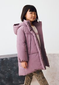 Lilla kapuutsiga polsterdatud mantel esiküljel luku ja neetidega. Modell kannab all valget särki ja leopardimustrilisi pükse.