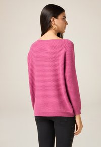 Różowy sweter z dzianiny o fakturowanym wzorze, z okrągłym dekoltem i rękawami do łokcia, zestawiony z czarnymi spodniami zdobionymi aplikacjami.