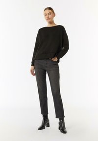 Czarna, oversize bluza z okrągłym dekoltem, zestawiona z ciemnymi dżinsami. Model ma na sobie czarne botki za kostkę i mało rzucający się w oczy design.