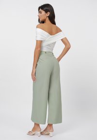 White off-shoulder top with twisted design, paired with high-waisted, wide-leg trousers in pale green. Model wears white heeled sandals.