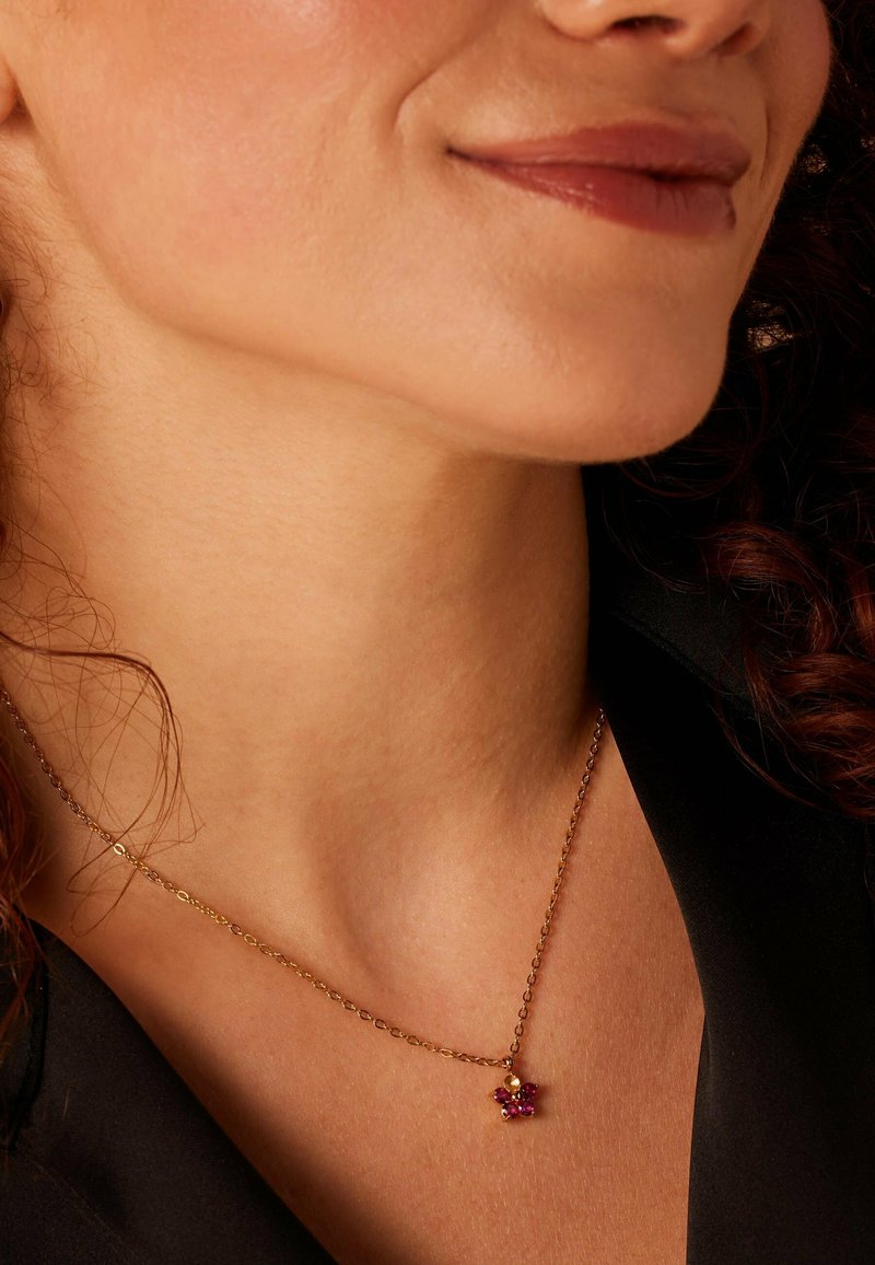 Gouden ketting met een bloemenvormige hanger versierd met rode edelstenen, rustend op de nek van een persoon met krullend haar.