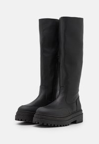 Mustat polvipituiset saappaat paksuilla urheilupohjilla ja sivuveskareilla, valmistettu sileästä nahasta, esillä valkoisella taustalla.