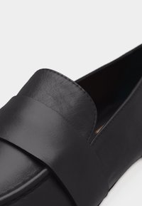 Μαύρο δερμάτινο μοκασίνι χωρίς κορδόνια με φαρδιά λεπτομέρεια ζώνης μπροστά και ορατή ραφή κατά μήκος των άκρων.