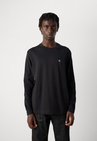 Ung mann med korte twists iført en svart langermet Calvin Klein-skjorte og mørke jeans, stående mot en enkel lys bakgrunn.