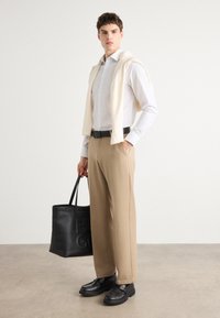 Hvit skjorte med lys beige genser drapert over skuldrene, brune bukser, svart belte og svart totebag. Svarte sko fullfører antrekket.