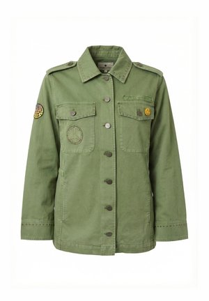 Giacca verde in stile militare con chiusura a bottoni, tasche sul petto, patch con il simbolo della pace, spilla con faccina sorridente e toppa ricamata con la scritta "Stay Weird".