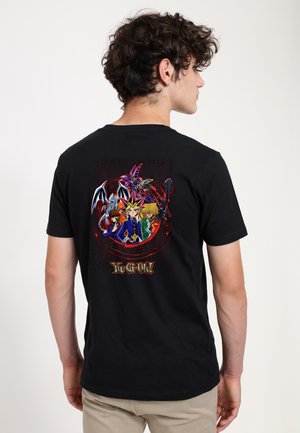 Ung mand med krøllet hår iført en sort t-shirt med farverige Yu-Gi-Oh!-karakterer og logo på bagsiden.