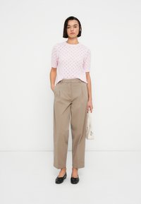 Ανοιχτό ροζ πλεκτό πουλόβερ με κοντά μανίκια, συνδυασμένο με μπεζ ψηλόμεσα παντελόνια. Το μοντέλο κρατάει μια μικρή λευκή τσάντα και φοράει μαύρα μπαλαρίνες.