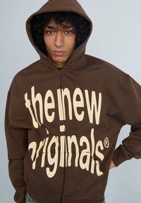 Smeđa zip-up dukserica s velikim kremastim natpisom "the new originals" sprijeda, s kapuljačom na vezanje i opuštenim krojem.