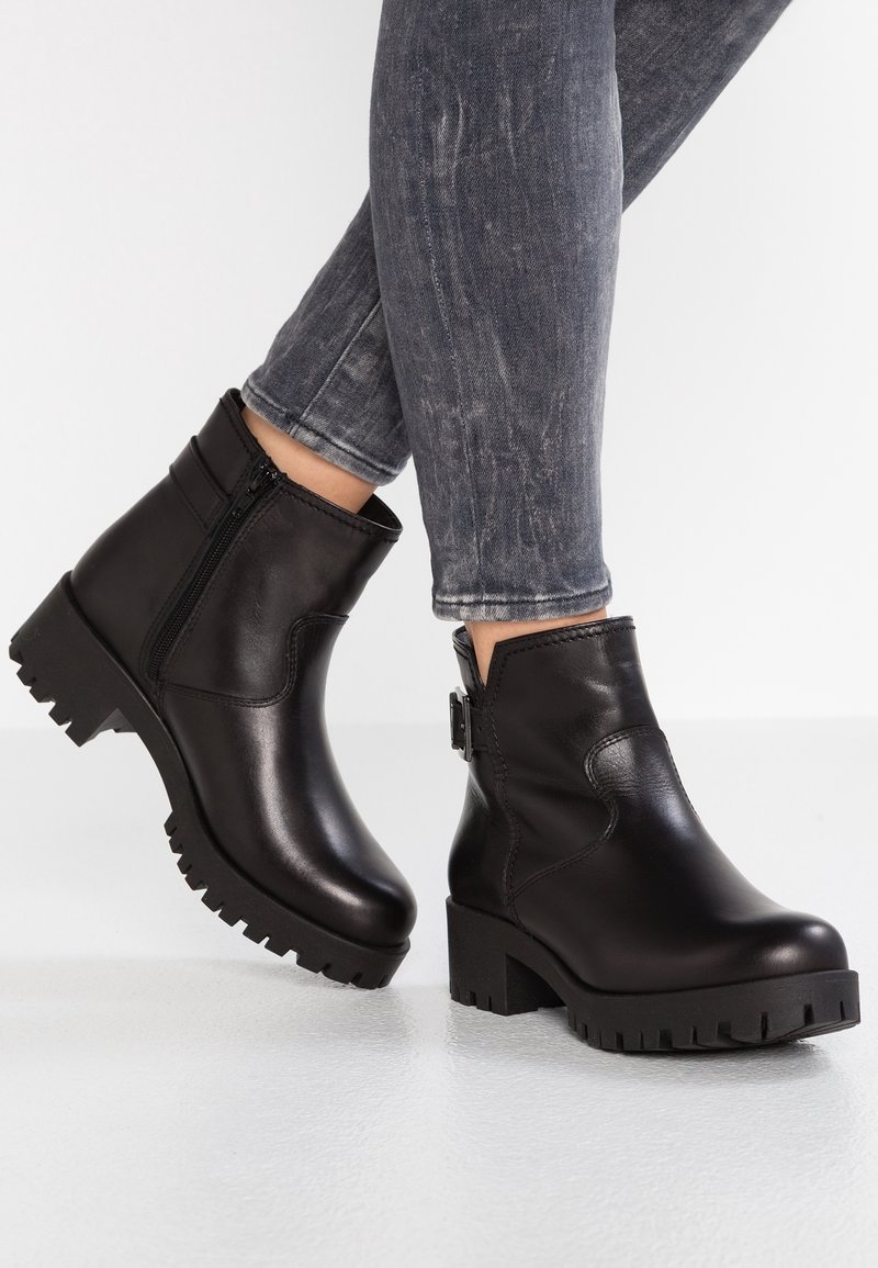 Paire de bottines en cuir noir à talons carrés et fermeture éclair latérale, portées avec un jean skinny gris délavé sur un pied levé.