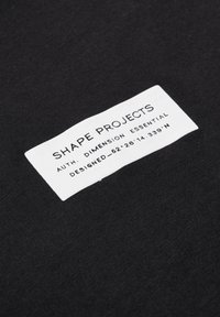 Svart tyg med en vit lapp. Lappen har text: "SHAPE PROJECTS," "AUTH. DIMENSION ESSENTIAL," och koordinater "DESIGNED - 52.26 14.339 N."