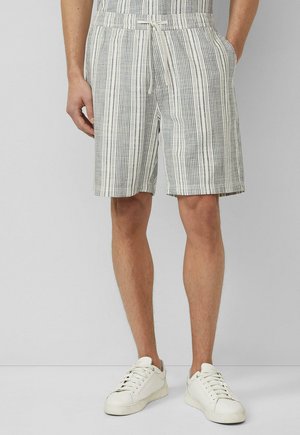 Mann trägt grau-weiß gestreifte Kordelzug-Shorts bis zur Mitte der Oberschenkel mit Seitentaschen und weißen Low-Top-Sneakern, steht auf hellem Boden.