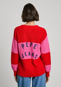 Adīta džemperis ar izteiksmīgām sarkanām un rozā horizontālām svītrām, ar "PEPE JEANS" melnā krāsā uz muguras. Ribota tekstūra un aproces.