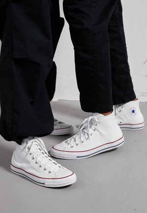 Due persone che indossano pantaloni neri e sneaker bianche alte Converse stanno in piedi su un pavimento grigio contro un muro bianco.