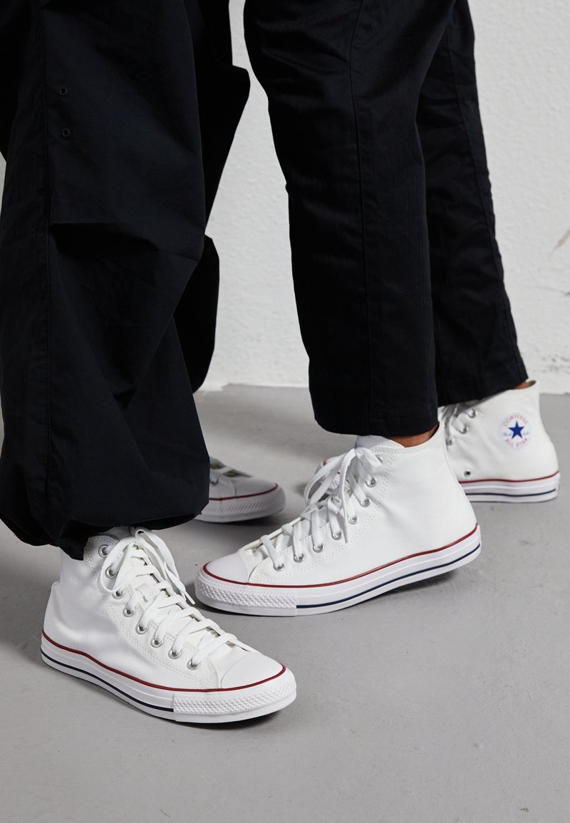 Deux personnes portant un pantalon noir et des baskets Converse blanches montantes se tiennent sur un sol gris contre un mur blanc.