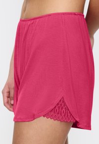 Różowe szorty z elastycznym pasem, z falbaniastym dołem i koronkowym wykończeniem, na górnej części uda i biodrze osoby na białym tle.