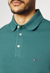 Polo shirt vert texturé avec trois boutons et un badge de marque sur la poitrine gauche. Présente un design de col classique.