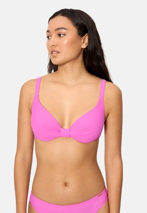 Mujer con cabello largo y oscuro, vestida con un bikini rosa brillante, mirando hacia su izquierda sobre un fondo claro y liso.