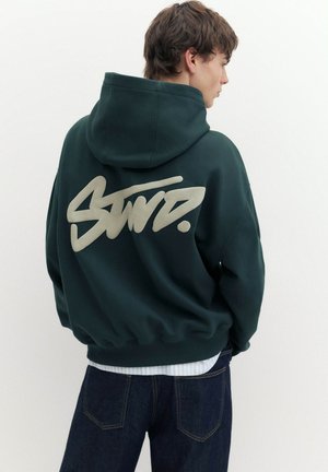 Junge Person, seitlich betrachtet, trägt einen dunkelgrünen Hoodie mit großen stilisierten weißen Buchstaben auf dem Rücken und dunkelblaue Jeans.