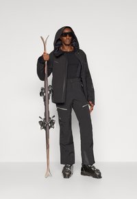 Die Skibekleidung besteht aus einer schwarzen wasserfesten Jacke mit Kapuze, schwarzen Hosen und Skischuhen. Skier mit Bindungen werden in einer Hand gehalten.