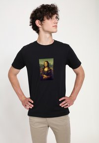 Černé bavlněné tričko s centrálním potiskem Mona Lisy. Model stojí se založenýma rukama v pase, aby ukázal design a střih.