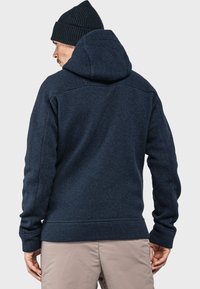 Marineblauer Fleece-Hoodie mit strukturierter Oberfläche, einer großen Kapuze und gerippten Bündchen, kombiniert mit neutral gefärbten Hosen und einer gestrickten Mütze.