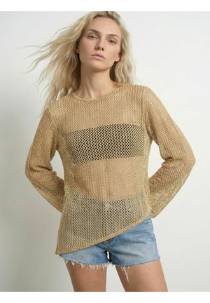Lyshåret kvinde iført beige åbenstrikket sweater over sort top og lyseblå denimshorts, står med hænderne bag ryggen.