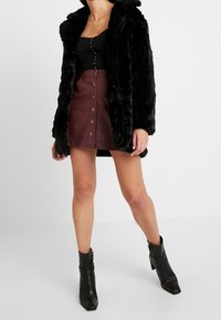 Musta tekoturkis takki, jossa on huppu, ribattu musta yläosa, viininpunainen nahkamenkkihame, jossa on napit, ja mustat nilkkurit, joissa on kärkiosa.