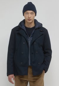 Cappotto navy doppiopetto con una superficie testurizzata, grandi bottoni e un rivestimento imbottito con cappuccio. Abbinato a un berretto a coste blu navy e un maglione scuro.