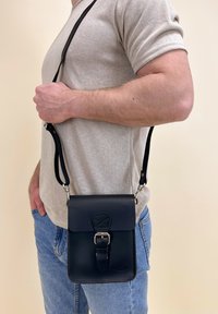 Schwarze Leder-Crossbody-Tasche mit Klappe, Metallverschluss und verstellbarem Tragegurt. Rechteckige Form mit glatter Textur und dezentem Logo.