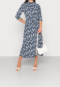 Mörkblå blommig midiklänning med korta ärmar, midjeband och vitt blommönster. Matchad med ljusa klacksandaler och en liten handväska.