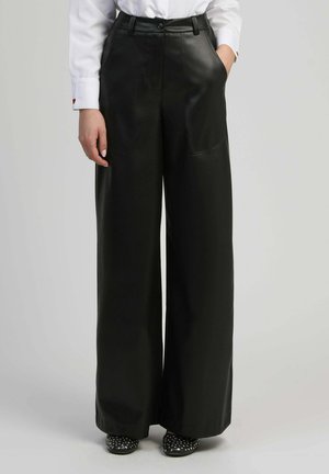 Personne portant un pantalon large en cuir noir taille haute avec poches avant, une chemise blanche avec un détail en forme de cœur sur le poignet, et des chaussures noires cloutées.