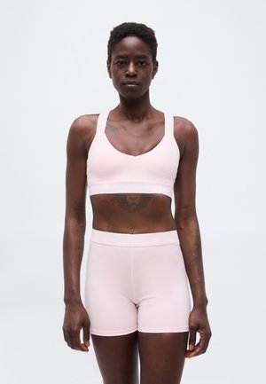 Frau mit kurzen lockigen Haaren, die einen hellrosa Sport-BH und passende Shorts trägt, vor einem schlichten weißen Hintergrund stehend.
