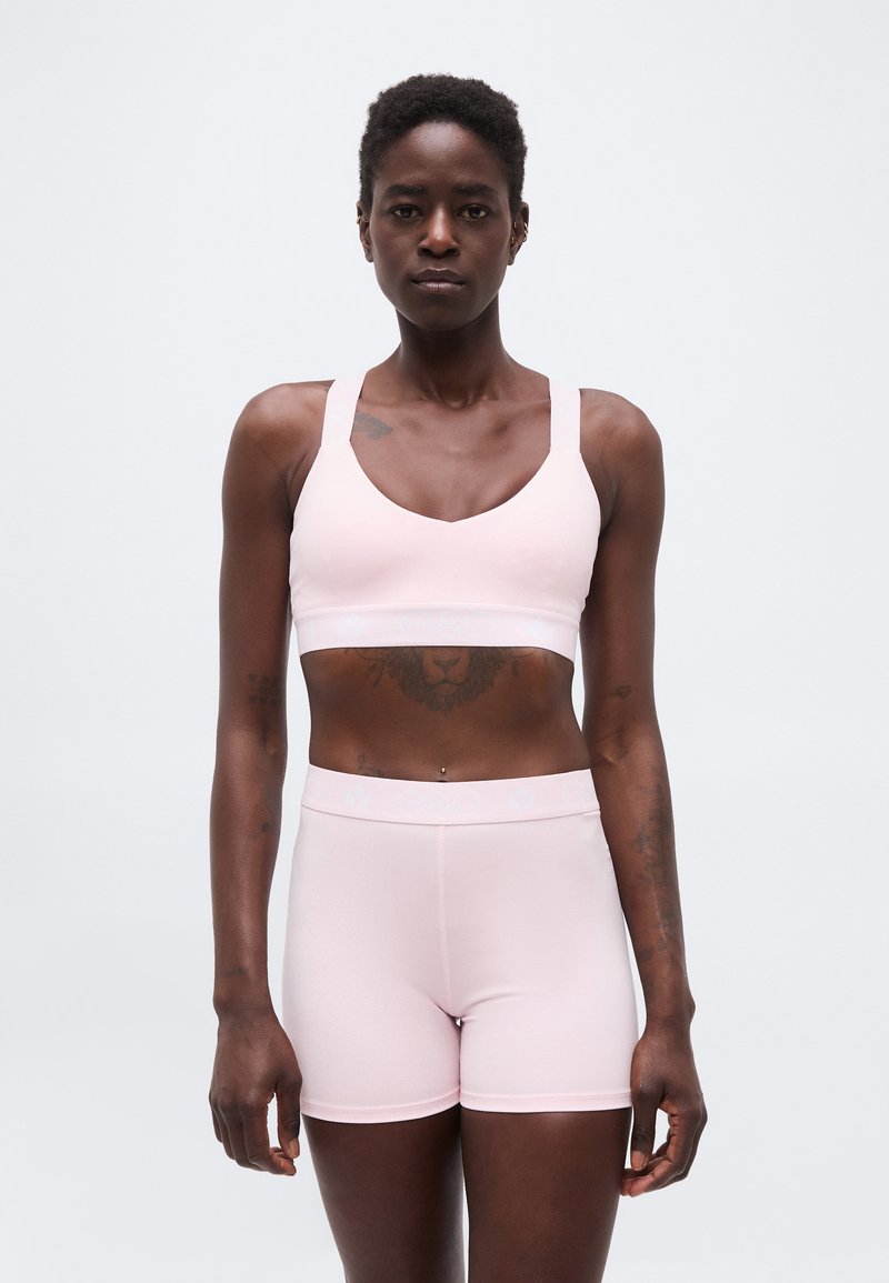 Γυναίκα με κοντά σγουρά μαλλιά, που φοράει αθλητικό μπουστάκι σε ανοιχτό ροζ χρώμα και αντίστοιχα σορτς, στέκεται μπροστά σε ένα απλό λευκό φόντο.