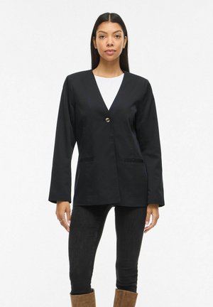 Kobieta ubrana w czarną marynarkę zapinaną na jeden guzik, białą bluzkę, czarne dżinsy i brązowe kozaki do połowy łydki, stojąca na tle jednolitego białego tła.