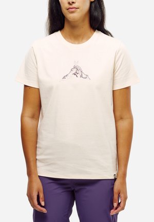 Femme portant un t-shirt beige clair avec un petit motif de montagne brodé au centre et un pantalon violet, debout devant un fond blanc.