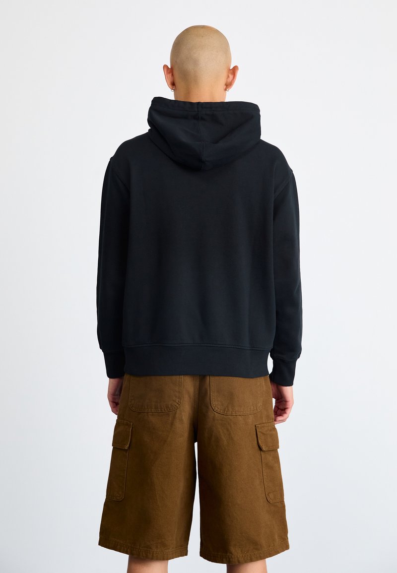 Felpa nera con cappuccio e bordo a coste, abbinata a pantaloni cargo marroni con tasche. Vista da dietro su uno sfondo neutro.