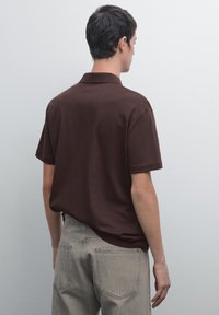 Hnědý polo tričko s krátkým rukávem a límečkem, hladký povrch a uvolněný střih, zkombinováno se světle šedými džíny. Model je viděn zezadu.