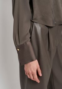 Bruine satijnen blouse met lange mouwen en een manchet met een gouden accent. De blouse is gecombineerd met bijpassende glanzende, bruine broek.