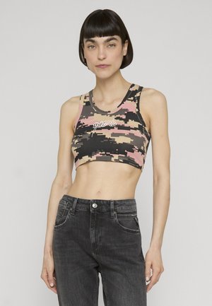 Vrouw met kort donker haar, die een roze, beige en zwarte digitale camo crop top draagt en hoge zwarte jeans voor een effen achtergrond.
