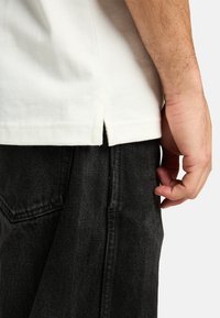 Camiseta blanca de algodón con aberturas laterales, combinada con jeans de mezclilla oscura. La tela parece suave, con una textura lisa y costuras mínimas.