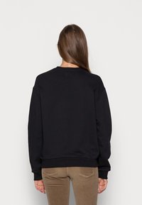 Sudadera negra de manga larga, con puños acanalados y cuello redondo. Textura de tela suave. Llevada sobre pantalones marrones.