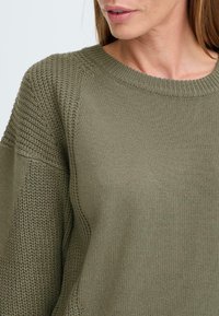 Olivgrön stickad tröja med rund halsringning, texturerade ärmar och avslappnad passform, med en blandning av släta och ribbade tygmönster.