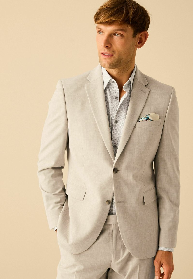 Costume gris clair avec une finition texturée, doté d'un revers cranté et de deux boutons. En dessous, une chemise bleu clair à motifs et un pochette.