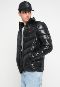 Juodas pūkinis džemperis su blizgiu paviršiumi, horizontalaus siuvimo dizainu, aukštu apykakle ir logotipu ant kairiojo rankogalių. Dėvimas ant pilkos spalvos marškinėlių.