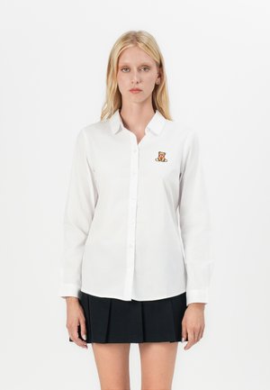 White long-sleeve button-up shirt with a small embroidered teddy bear on the chest, paired with a pleated black skirt.