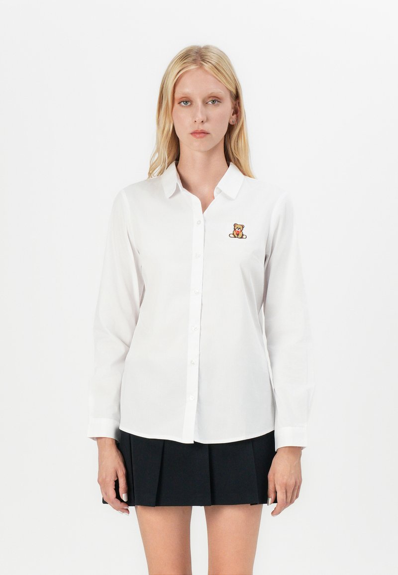 White long-sleeve button-up shirt with a small embroidered teddy bear on the chest, paired with a pleated black skirt.